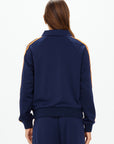 A person with long brown hair, seen from behind, wears The Upside's VITESSE PARI ZIP JACKET in navy with orange sleeve stripes and matching slinky pants, set against a plain white background.