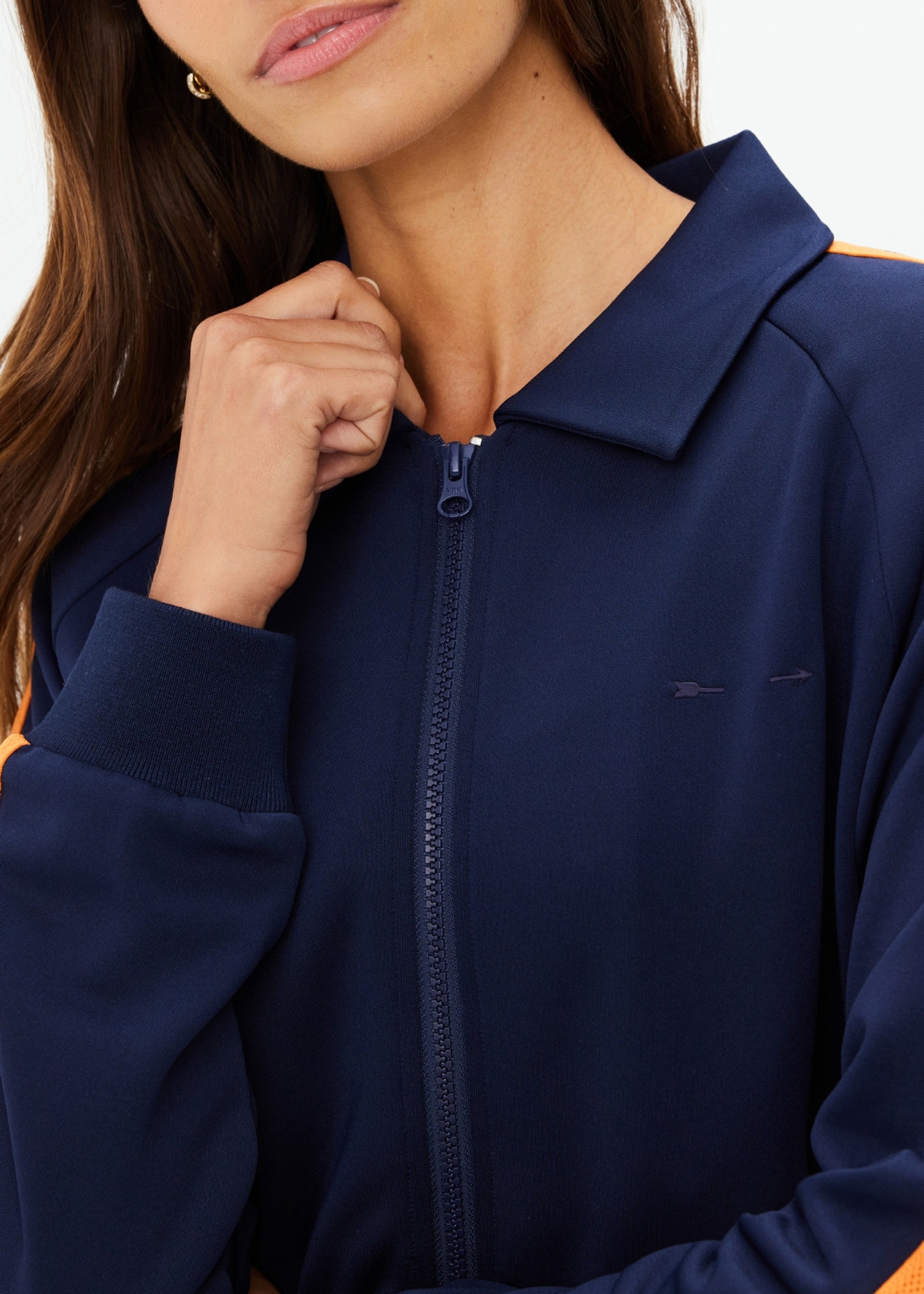A woman in The Upside's VITESSE PARI ZIP JACKET—navy blue with orange sleeve stripes and collar—is shown from the shoulders up, holding the zipper near her chin. Her face is partially visible. Pairs well with matching pants.