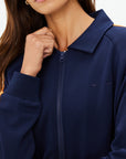 A woman in The Upside's VITESSE PARI ZIP JACKET—navy blue with orange sleeve stripes and collar—is shown from the shoulders up, holding the zipper near her chin. Her face is partially visible. Pairs well with matching pants.
