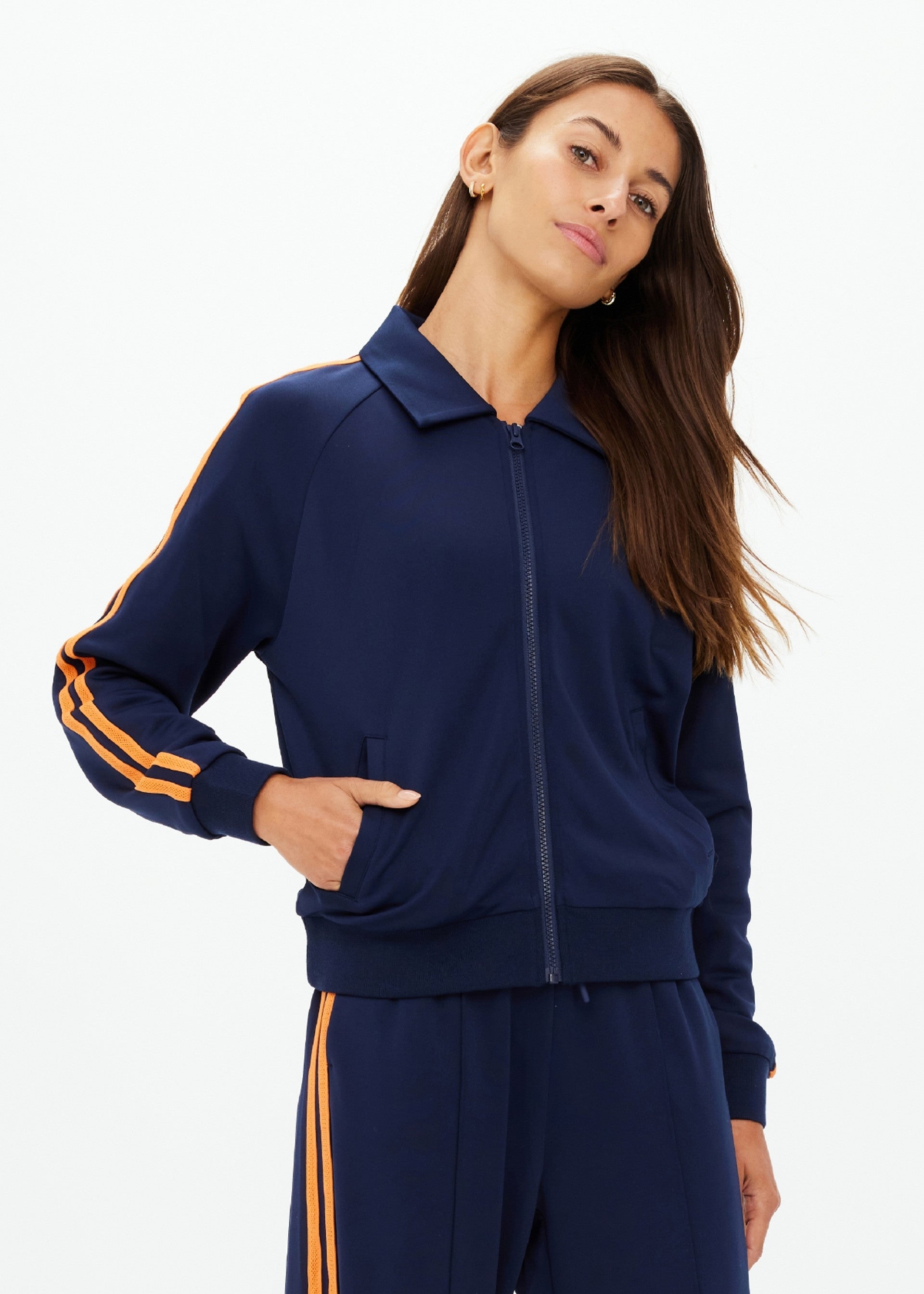 A woman wears The Upside’s VITESSE PARI ZIP JACKET in navy blue with orange stripes and matching slinky pants, standing with one hand in her pocket and looking to the side.