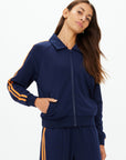 A woman wears The Upside’s VITESSE PARI ZIP JACKET in navy blue with orange stripes and matching slinky pants, standing with one hand in her pocket and looking to the side.