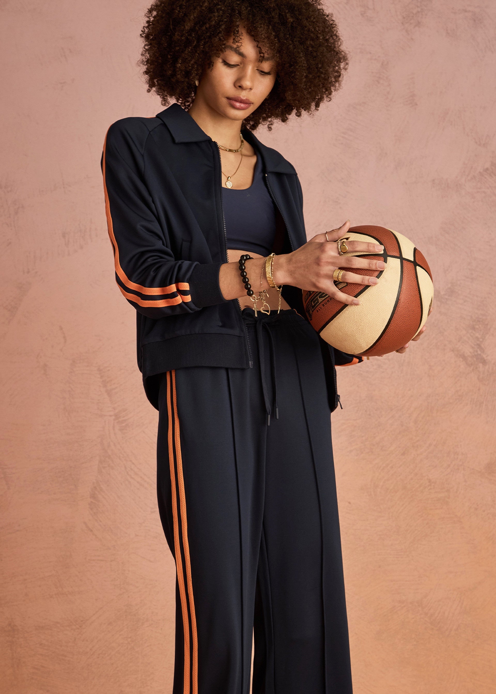 A curly-haired woman stands against a textured pink background wearing The Upside’s VITESSE PARI ZIP JACKET, a navy blue sports jacket with orange stripes made from recycled PE, and holds a basketball in her hands.