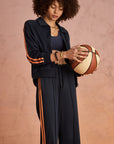 A curly-haired woman stands against a textured pink background wearing The Upside’s VITESSE PARI ZIP JACKET, a navy blue sports jacket with orange stripes made from recycled PE, and holds a basketball in her hands.