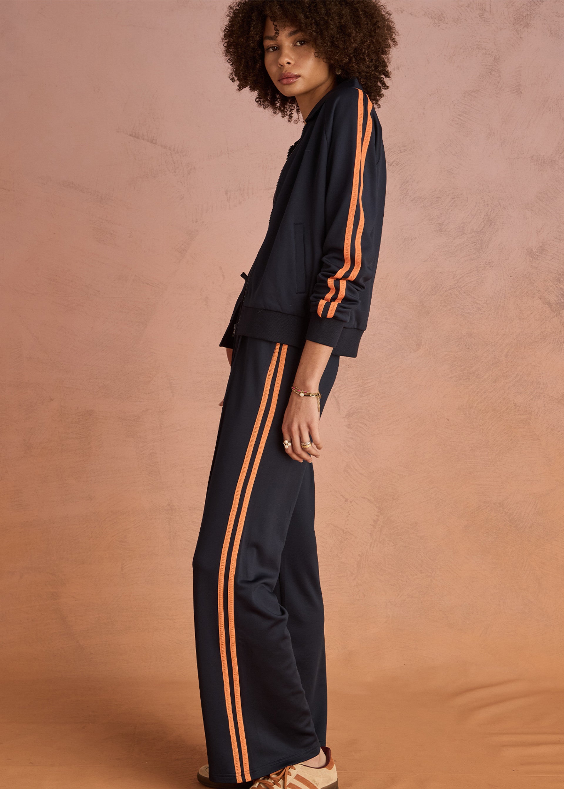 A person with curly hair stands sideways against a peach background, wearing The Upside’s VITESSE PARI ZIP JACKET in navy with orange stripes and matching pants in slinky fabric, paired with beige sneakers.