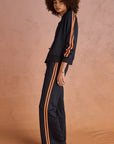 A person with curly hair stands sideways against a peach background, wearing The Upside’s VITESSE PARI ZIP JACKET in navy with orange stripes and matching pants in slinky fabric, paired with beige sneakers.