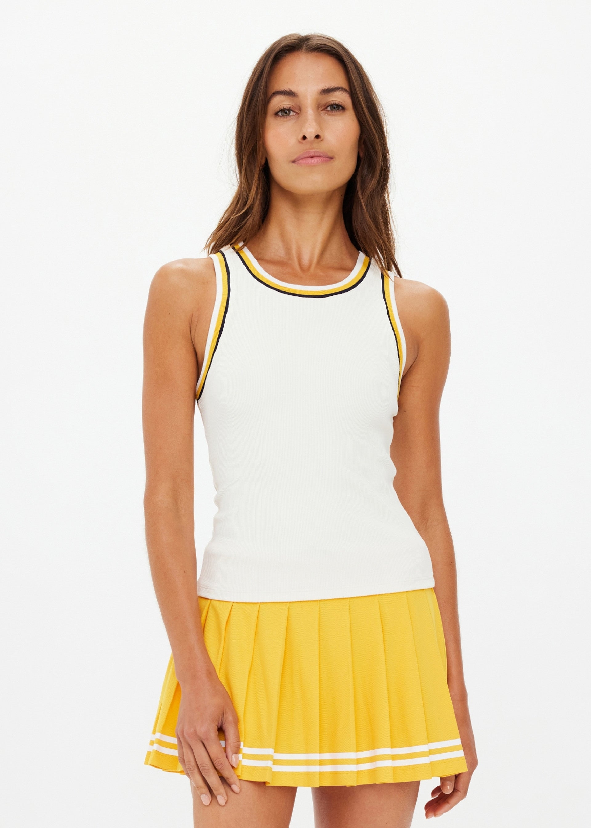 A woman faces forward against a white background, wearing The Upside's KIRSTY RIB TANK TOP with black and yellow trim, paired with a yellow pleated skirt featuring white stripes near the hem.