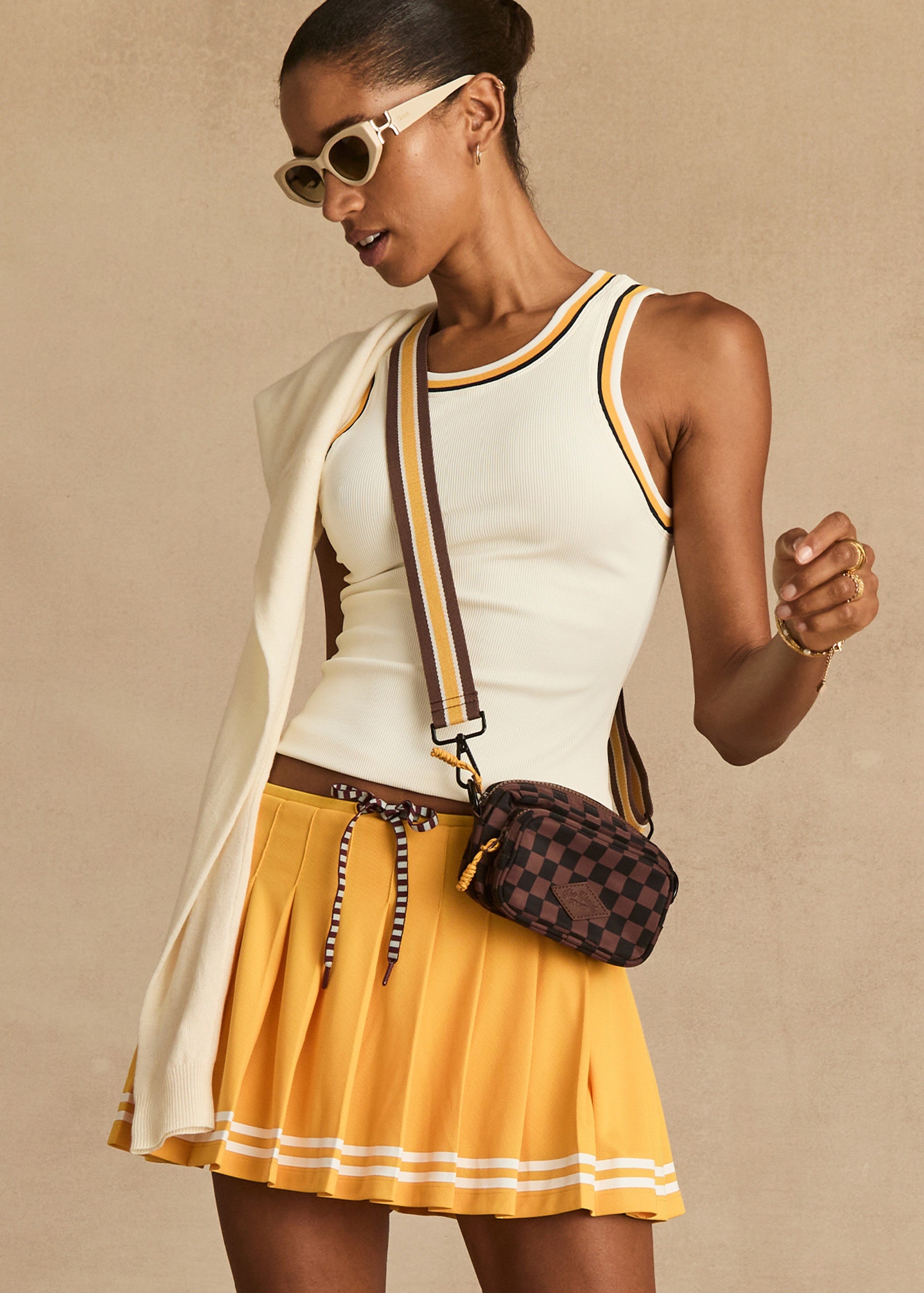 A woman wearing The Upside’s KIRSTY RIB TANK TOP, white sunglasses, a yellow pleated skirt, and a checkered crossbody bag with a sweater over her shoulder stands against a beige background.