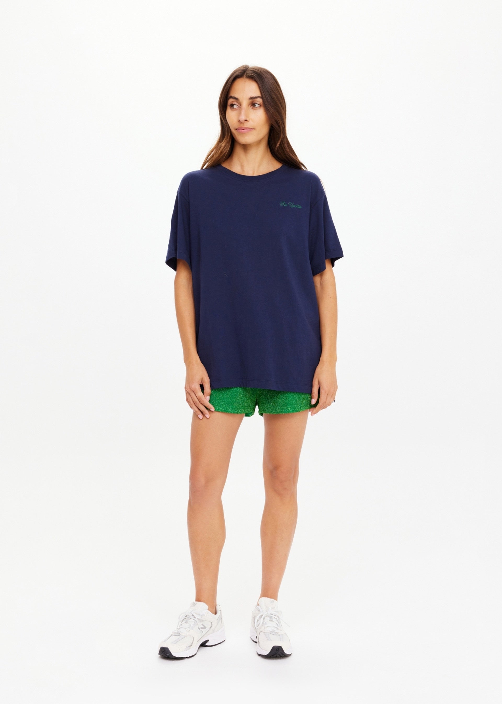 A woman with long brown hair and a neutral expression faces forward against a white background, wearing The Upside's SAMMI TEE in navy blue, paired with green shorts and white sneakers.