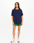 A woman with long brown hair and a neutral expression faces forward against a white background, wearing The Upside's SAMMI TEE in navy blue, paired with green shorts and white sneakers.