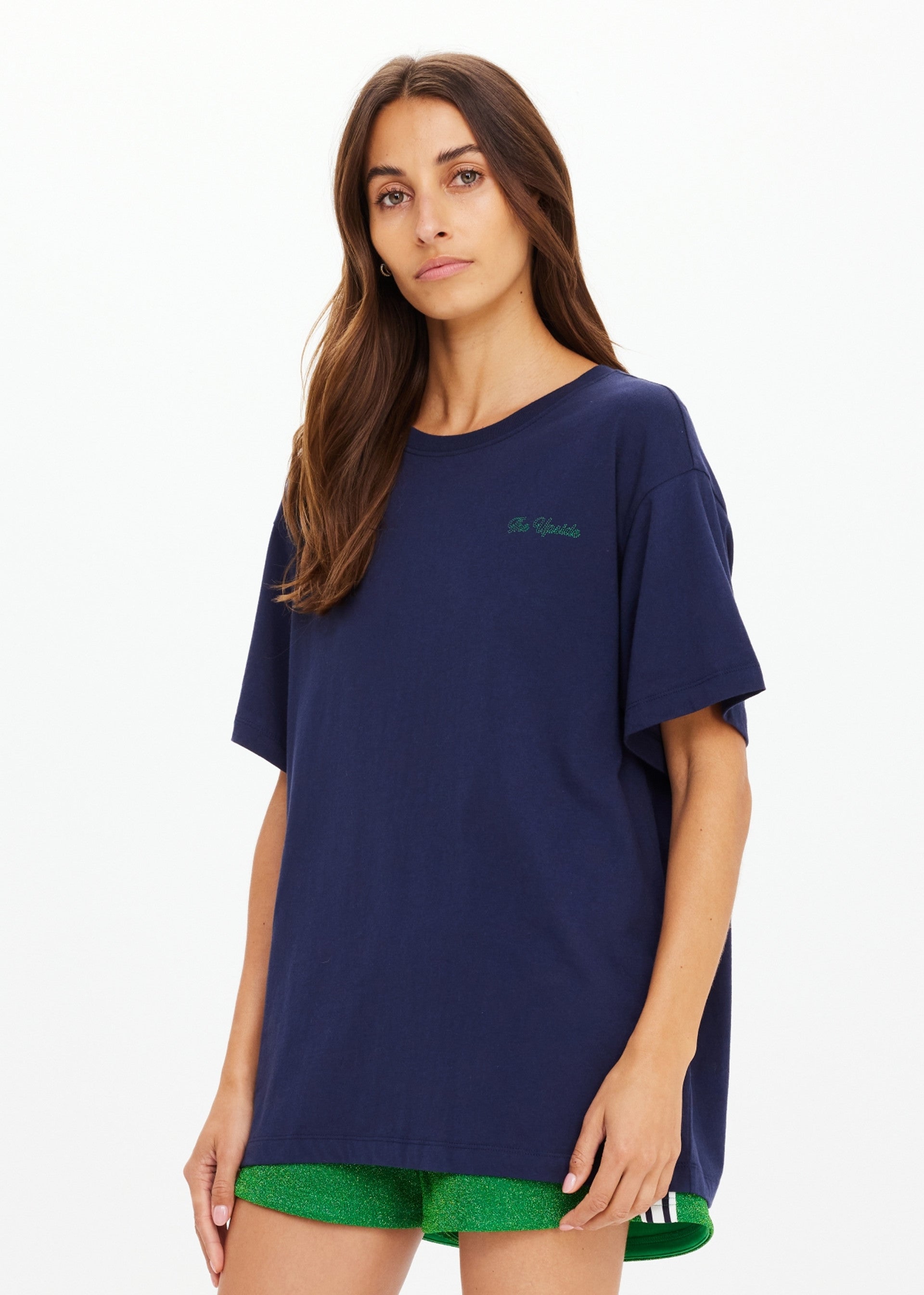 A woman with long brown hair wears The Upside SAMMI TEE and bright green shorts, standing against a plain white background and looking at the camera with a neutral expression.