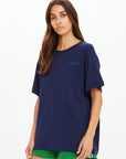 A woman with long brown hair wears The Upside SAMMI TEE and bright green shorts, standing against a plain white background and looking at the camera with a neutral expression.