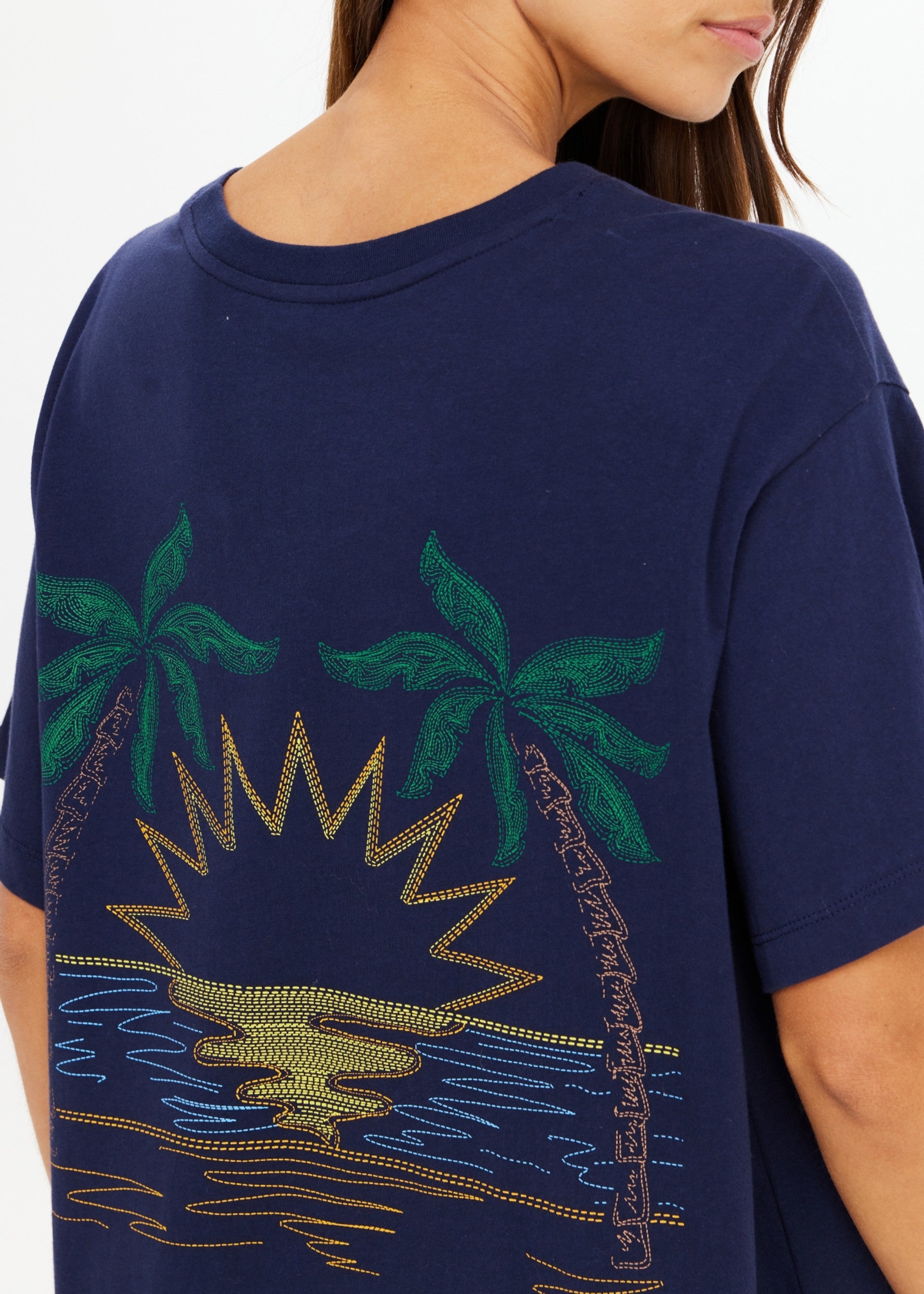 A woman wears The Upside’s SAMMI TEE, an oversized navy graphic tee with an embroidered palm tree, sunset, and water design on the back. Only her back and part of her face are visible.