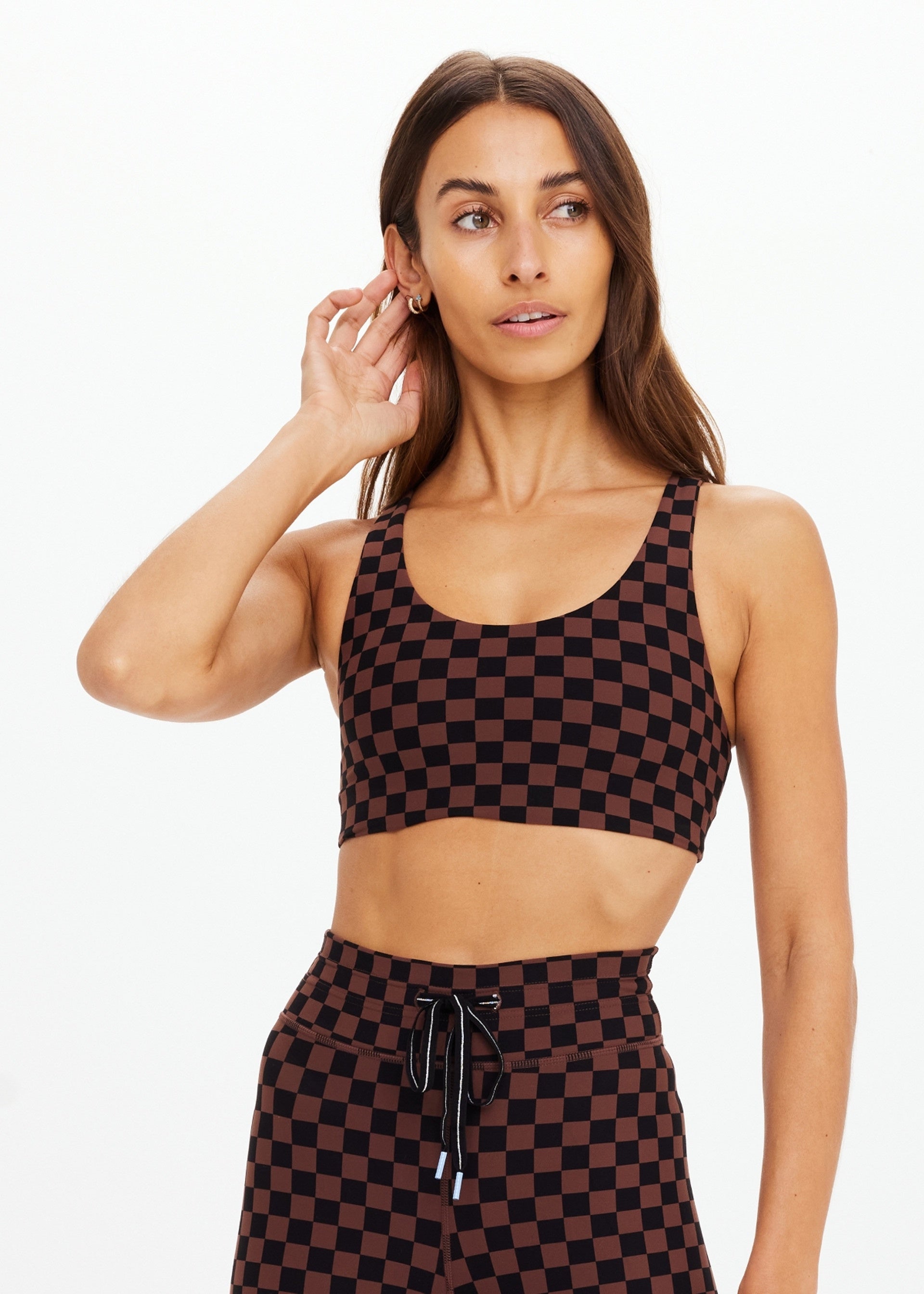 A woman wears The Upside TECHNICA JADE BRA with matching high-waisted leggings in brown and black checks, standing against a plain white background, looking left with one hand touching her hair.