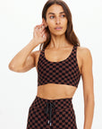 A woman wears The Upside TECHNICA JADE BRA with matching high-waisted leggings in brown and black checks, standing against a plain white background, looking left with one hand touching her hair.