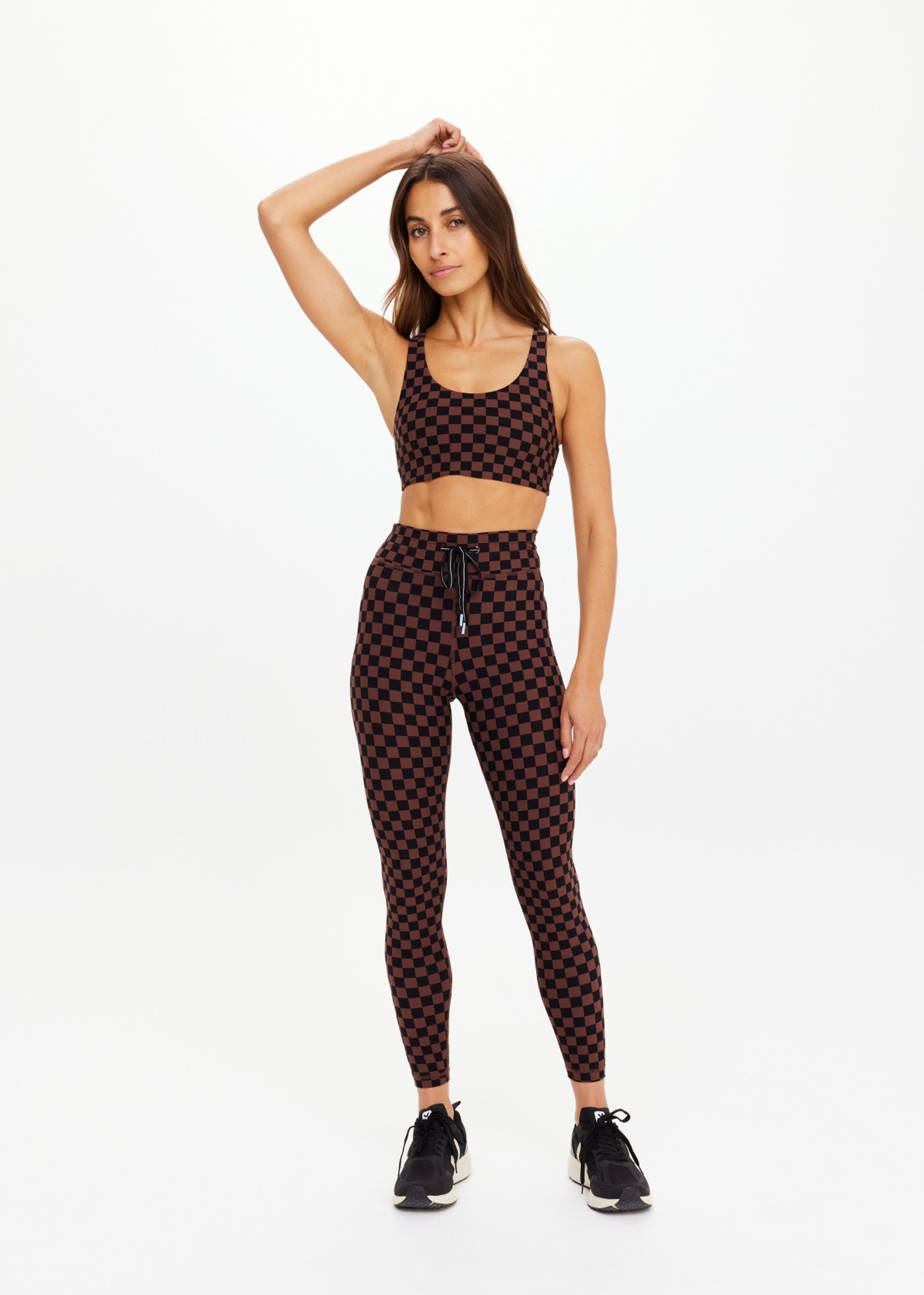 A woman poses in The Upside TECHNICA JADE BRA, a moisture-wicking brown and black checkered sports bra, paired with matching leggings and black sneakers. She stands with one hand on her head against a plain white background.