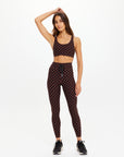 A woman poses in The Upside TECHNICA JADE BRA, a moisture-wicking brown and black checkered sports bra, paired with matching leggings and black sneakers. She stands with one hand on her head against a plain white background.