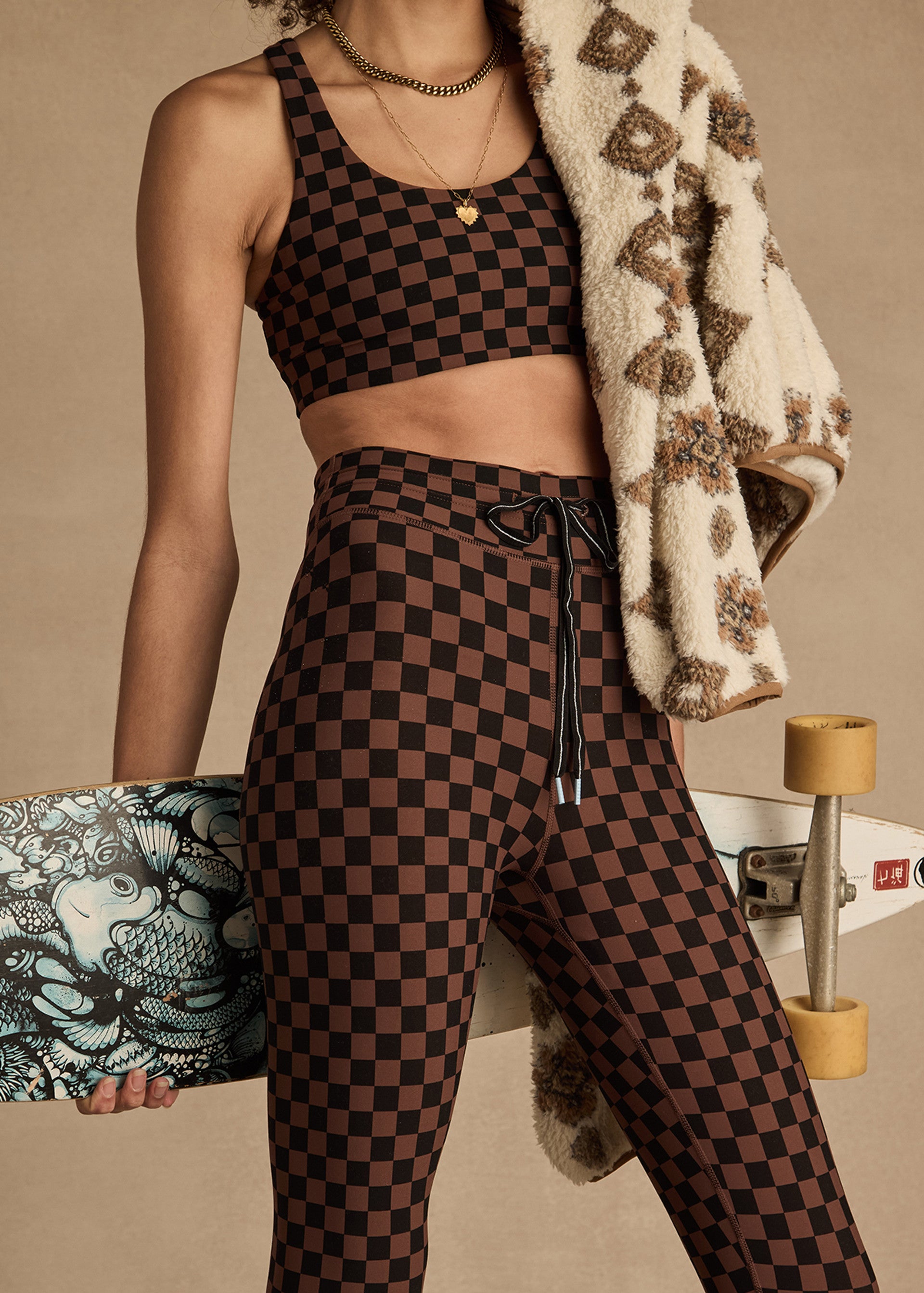 Wearing The Upside’s TECHNICA JADE BRA in a brown and black checkerboard print with matching leggings, a person holds a blue skateboard and drapes a patterned fleece jacket over their shoulder. Their face is not shown.