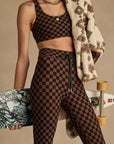 Wearing The Upside’s TECHNICA JADE BRA in a brown and black checkerboard print with matching leggings, a person holds a blue skateboard and drapes a patterned fleece jacket over their shoulder. Their face is not shown.