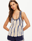 A woman with long brown hair is wearing a sleeveless WISHBONE EMMA KNITTED TANK, which features a hand tie-dyed blue pattern on beige fabric. She pairs it with black athletic shorts that have white trim, and her arms are resting by her sides as she stands against a plain light background.