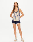 A person with long hair stands confidently, wearing the WISHBONE EMMA KNITTED TANK featuring a blue and white hand tie-dyed design crafted from organic cotton. They pair it with dark blue athletic shorts accented with white trim and complete the look with white sneakers. The scene is set against a plain white studio backdrop.