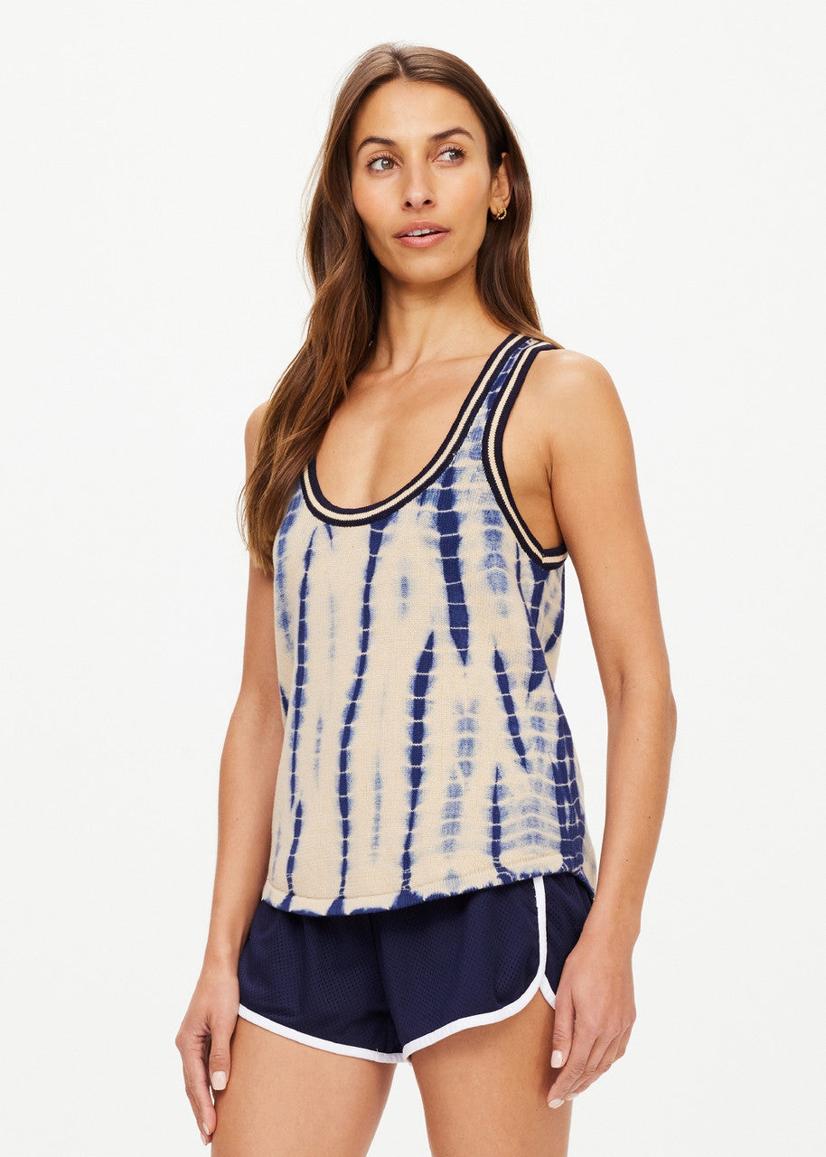 A woman with long brown hair is wearing the WISHBONE EMMA KNITTED TANK, a sleeveless, hand tie-dyed top made from organic cotton. She pairs it with navy athletic shorts featuring white trim, standing against a plain white background and gazing slightly to the side.