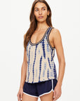 A woman with long brown hair is wearing the WISHBONE EMMA KNITTED TANK, a sleeveless, hand tie-dyed top made from organic cotton. She pairs it with navy athletic shorts featuring white trim, standing against a plain white background and gazing slightly to the side.