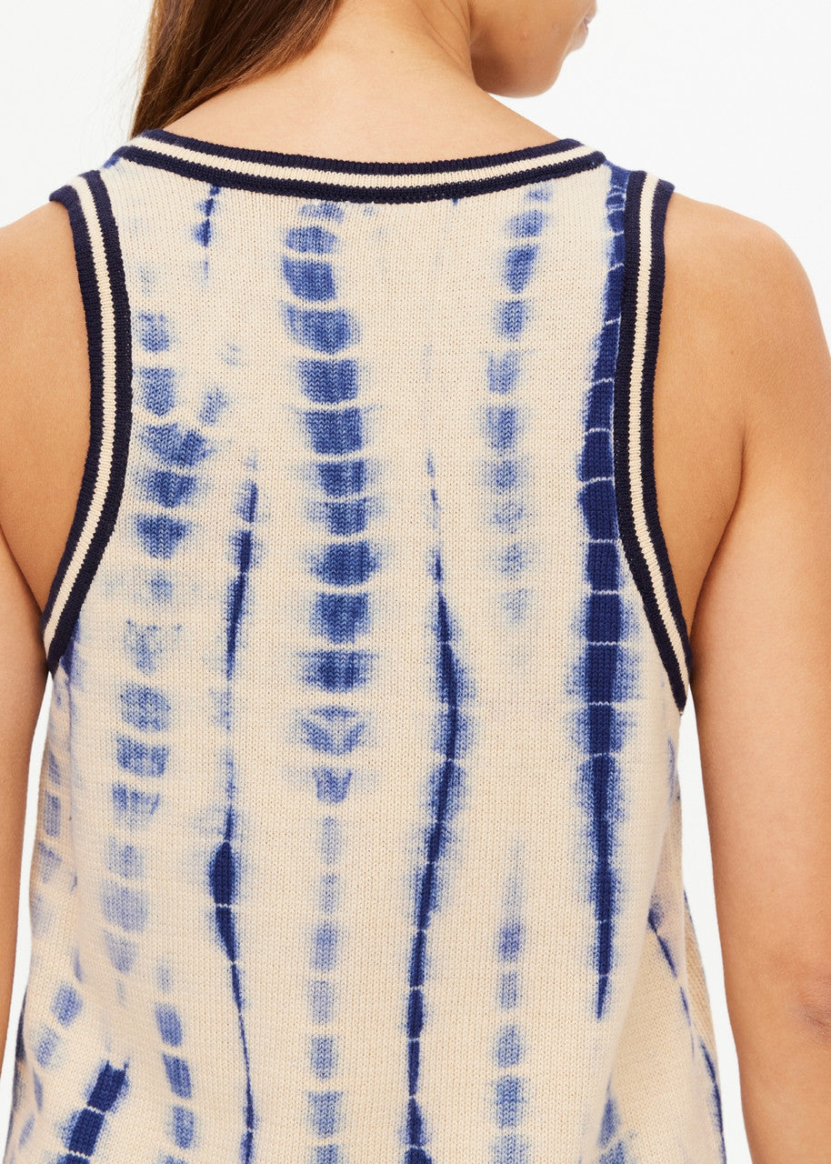 A close-up shot highlights the back of an individual wearing the WISHBONE EMMA KNITTED TANK, a sleeveless top featuring a hand tie-dyed design in beige with blue vertical streaks. The tank is accented with dark trim on the armholes and neckline. The person, with long brown hair, has their head turned away from the camera.