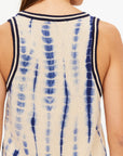 A close-up shot highlights the back of an individual wearing the WISHBONE EMMA KNITTED TANK, a sleeveless top featuring a hand tie-dyed design in beige with blue vertical streaks. The tank is accented with dark trim on the armholes and neckline. The person, with long brown hair, has their head turned away from the camera.