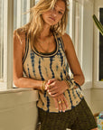 A woman with wavy blonde hair stands leaning against a wall in a sunlit room. She wears the beautifully crafted WISHBONE EMMA KNITTED TANK, patterned dark green shorts, and layered necklaces and bracelets. A cactus sits near the window beside her.