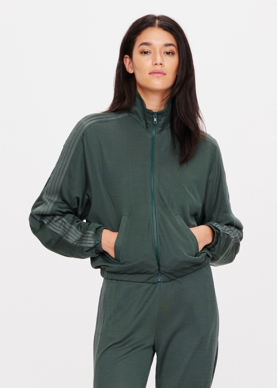 A person wearing The Upside's Roulette Lopez Jacket in deep green, paired with matching pants, stands against a white background. The cropped zip-through jacket features striped detailing on the shoulders and sleeves. With hands in the jacket pockets, the ensemble exudes casual confidence.