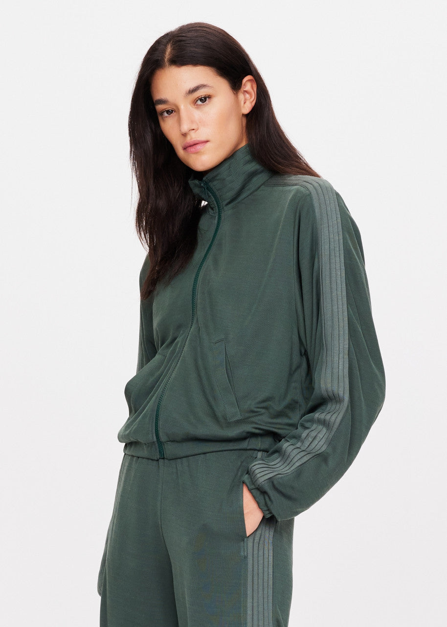 A person with long dark hair wears The Upside's Roulette Lopez Jacket, which comes in a deep green color and features stripes on the sleeves. With their right hand tucked into their pocket, they stand against a plain background, exuding casual confidence.