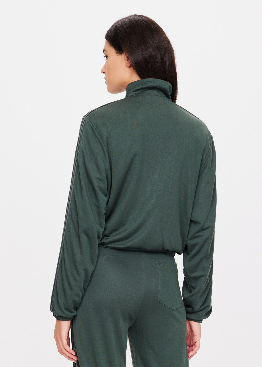 A person with long dark hair is facing away from the camera, wearing The Upside's Roulette Lopez Jacket in a deep green color with a high collar. The jacket, featuring blouson sleeves and subtly visible stripes on the sleeves, stands out against the plain, light-colored background.