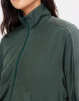 A person wearing The Upside's Roulette Lopez Jacket, a dark green zip-up with a high collar. The deep green fabric showcases a subtle striped pattern on the shoulder, and the face is partially out of the frame.