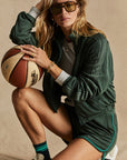 A person wearing The Upside's Roulette Lopez Jacket in a deep green fabric, along with matching shorts and sunglasses, crouches while holding a basketball. With long hair and sneakers complemented by striped socks, they strike a pose against a plain background.