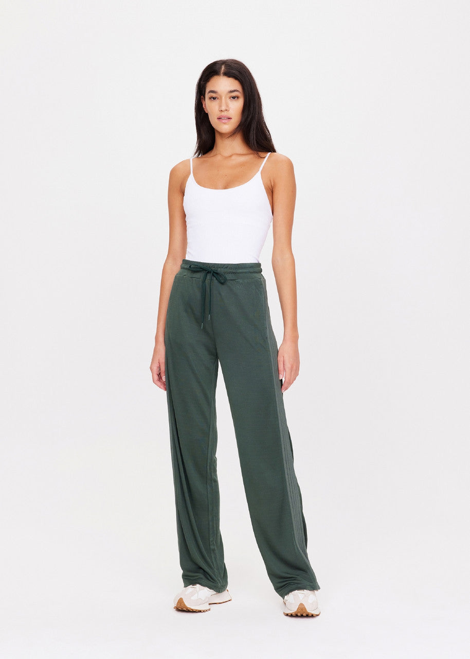 A woman in a white tank top paired with the Roulette Honour Pant from The Upside, featuring high-rise wide-leg design and slinky deep green fabric with an elasticated waist.