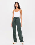 A woman in a white tank top paired with the Roulette Honour Pant from The Upside, featuring high-rise wide-leg design and slinky deep green fabric with an elasticated waist.