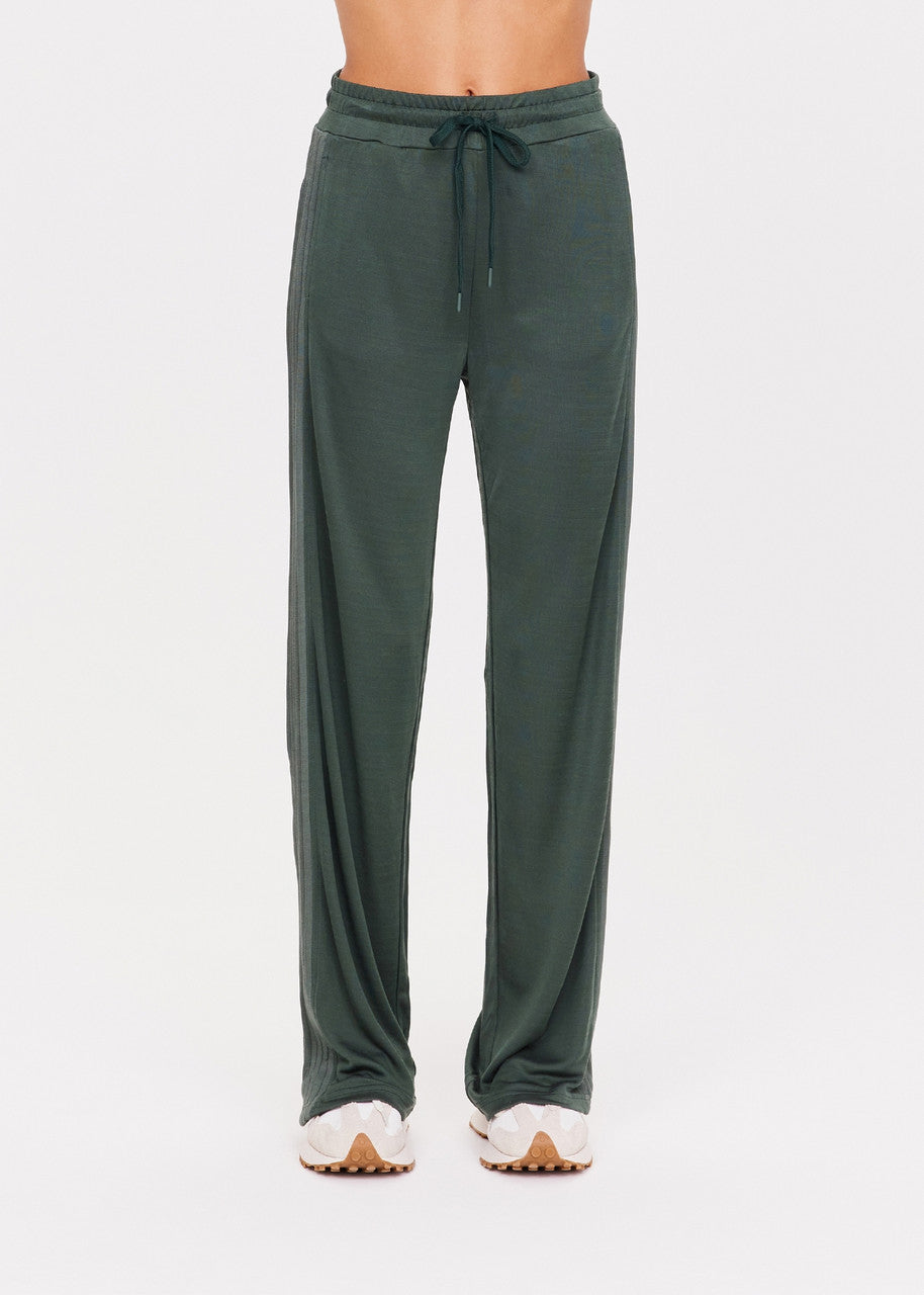 A person wearing The Upside's Roulette Honour Pant in dark green, featuring a high-rise wide-leg design with an elasticated waist and a drawstring. The slinky deep green fabric adds a casual flair, perfectly paired with white sneakers against a plain white background.