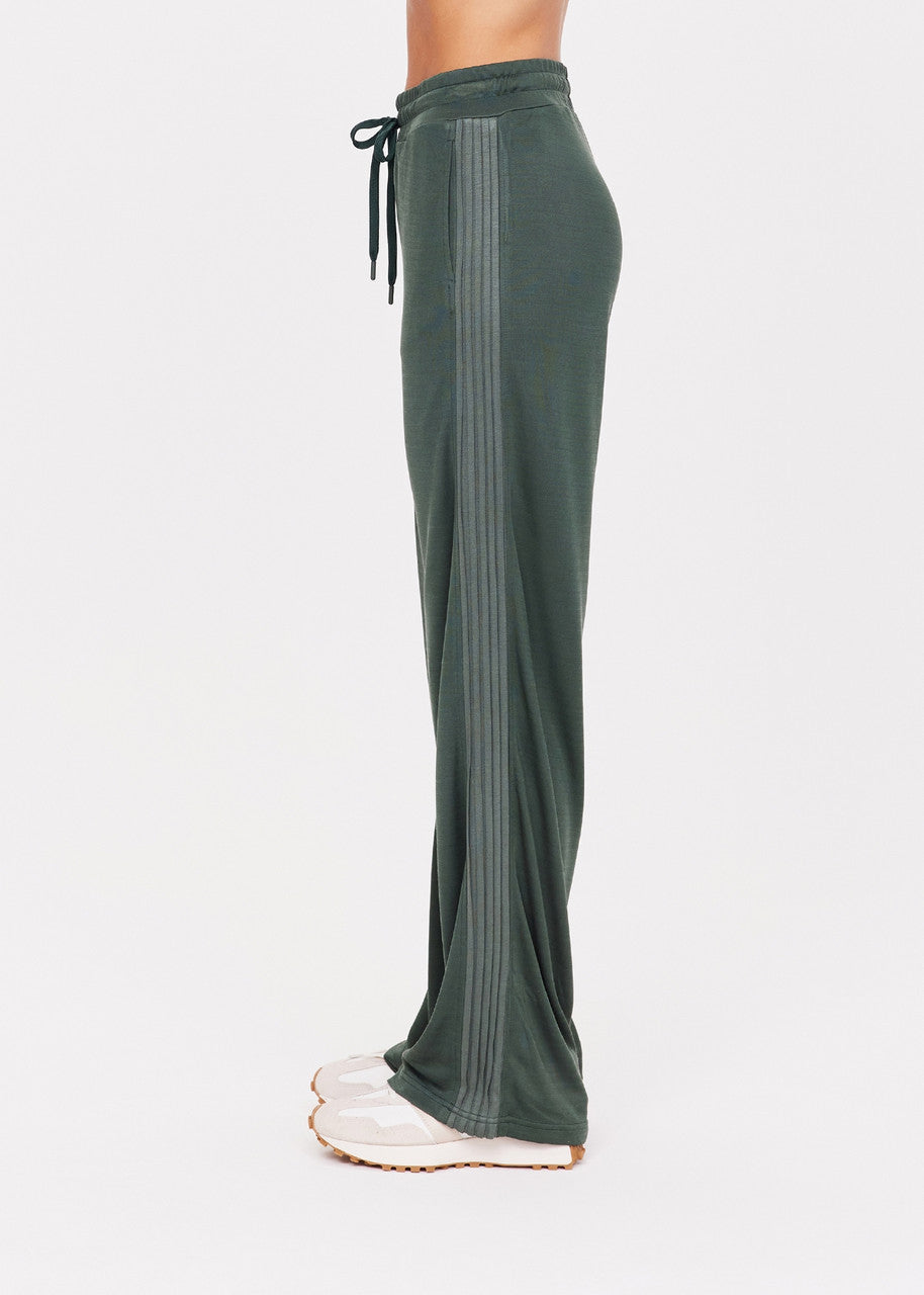 A person stands in profile wearing The Upside's Roulette Honour Pant, which features a high-rise, wide-leg silhouette made of slinky deep green fabric with side stripes and an elasticated drawstring waist, paired with white shoes. The background is plain white.