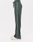 A person stands in profile wearing The Upside's Roulette Honour Pant, which features a high-rise, wide-leg silhouette made of slinky deep green fabric with side stripes and an elasticated drawstring waist, paired with white shoes. The background is plain white.