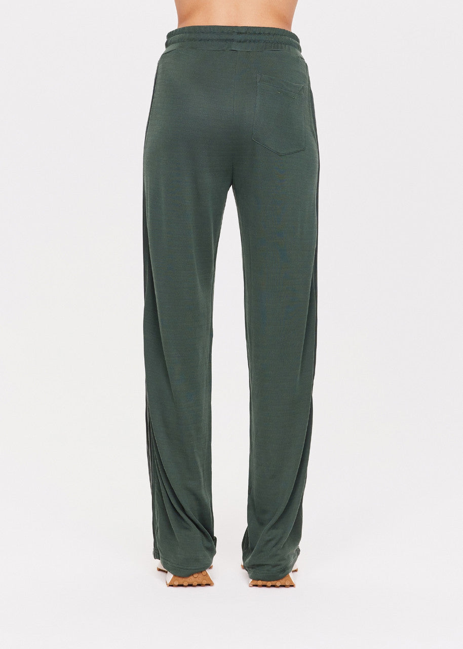 A person is wearing The Upside's Roulette Honour Pant, which features a high-rise design with an elasticated waist and is made from luxurious slinky deep green fabric. These wide-leg pants have a loose fit, visible back pocket, and reach the floor. They are styled with brown sandals against a plain white background.