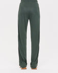 A person is wearing The Upside's Roulette Honour Pant, which features a high-rise design with an elasticated waist and is made from luxurious slinky deep green fabric. These wide-leg pants have a loose fit, visible back pocket, and reach the floor. They are styled with brown sandals against a plain white background.