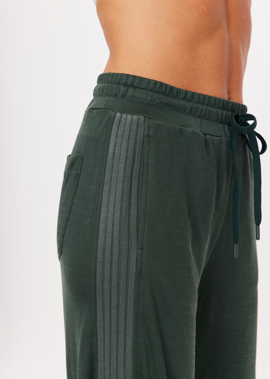 A close-up of a person wearing The Upside's Roulette Honour Pant, which are dark green athletic sweatpants featuring drawstrings and an elasticated waist. The pants showcase a textured side stripe design in slinky deep green fabric, emphasizing the fit and material around the waist and upper legs.