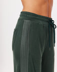 A close-up of a person wearing The Upside's Roulette Honour Pant, which are dark green athletic sweatpants featuring drawstrings and an elasticated waist. The pants showcase a textured side stripe design in slinky deep green fabric, emphasizing the fit and material around the waist and upper legs.