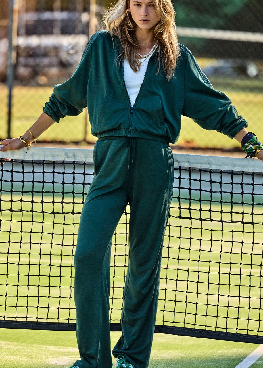 Wearing the Roulette Honour Pant by The Upside, a person stands on a tennis court in a fashionable ensemble, featuring deep green fabric with an elasticated waist over a white top. They lean casually against the net while tennis balls are scattered across the ground and a fence provides the backdrop.