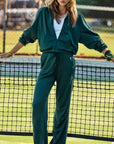 Wearing the Roulette Honour Pant by The Upside, a person stands on a tennis court in a fashionable ensemble, featuring deep green fabric with an elasticated waist over a white top. They lean casually against the net while tennis balls are scattered across the ground and a fence provides the backdrop.