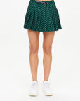 A person showcases The Upside's Ace of Hearts Cadiz Skirt, featuring a green pleated design with an elastic waistband and white polka dots, perfectly paired with crisp white sneakers. The scene is set against a plain white background.