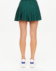 Rear view of a person wearing The Upside's Ace of Hearts Cadiz Skirt, a green polka-dotted, pleated design crafted from recycled polyamide, paired with white sneakers accented in red and blue, against a plain background.