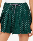 A person is wearing The Upside's Ace of Hearts Cadiz Skirt, which features a green pleated design with a white diamond pattern and a striped waist tie made from recycled polyamide. The individual complements the stylish skirt with a white top, emphasizing the chic look from the waist down.