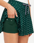 A person is wearing The Upside's Ace of Hearts Cadiz Skirt, featuring a green polka dot design with a striped drawstring. As they lift the fabric, you can see the built-in shorts with a pocket, made from recycled polyamide for an eco-friendly touch.