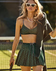 A woman wearing The Upside's Ace of Hearts Cadiz Skirt and a polka dot bikini top holds a tennis racket. She has a sweater draped over her shoulder, wears sunglasses and layered necklaces, standing against the backdrop of a tennis court that perfectly complements her recycled polyester ensemble.